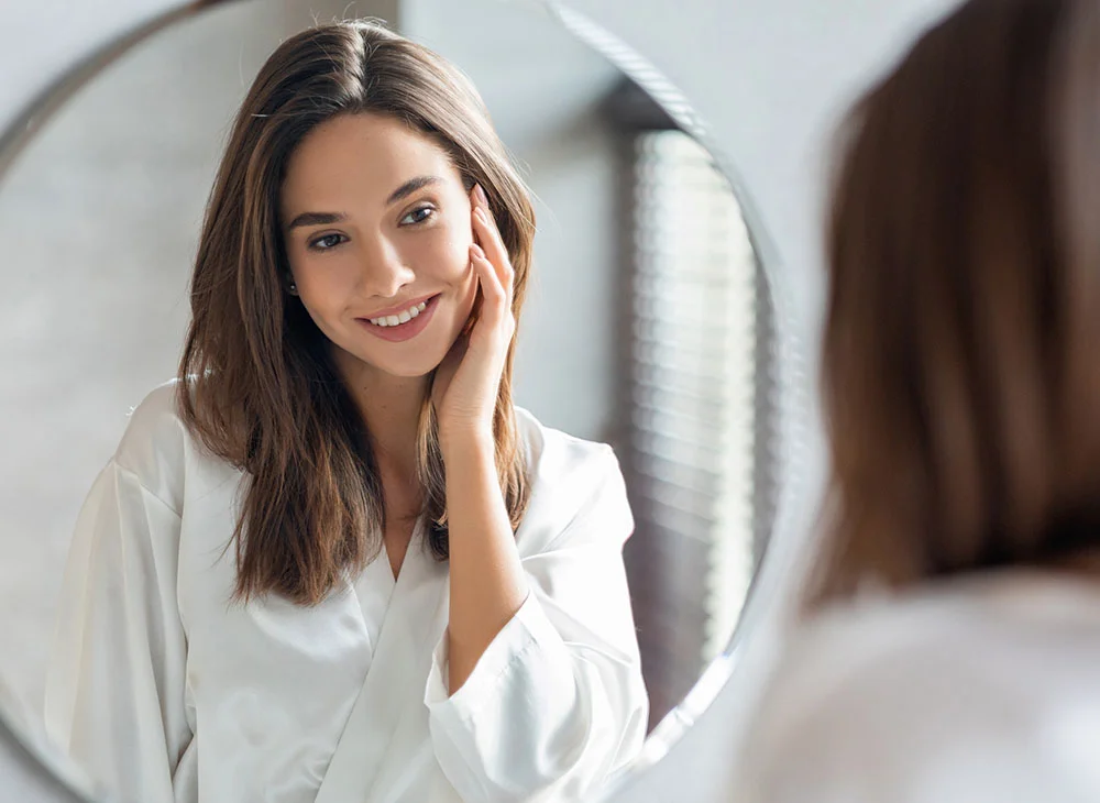 A woman with long brown hair wearing a white shirt looks at a mirror, with her reflection visible. She smiles gently while grazing her hand on her face, appearing relaxed and content. - Morpheus 8 in Aventura, FL