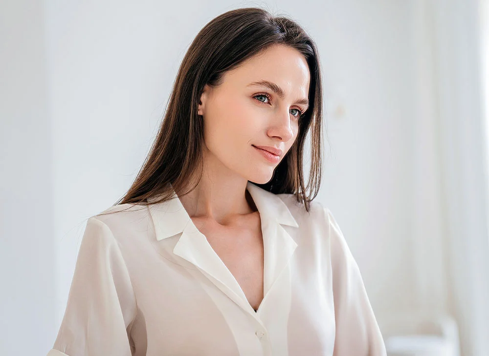 A woman in a beige blouse stands against a white background, smiling as she faces slightly to the side of the camera. She has long brown hair, exuding a friendly and approachable demeanor. - Non-Surgical Browlift in Aventura, FL