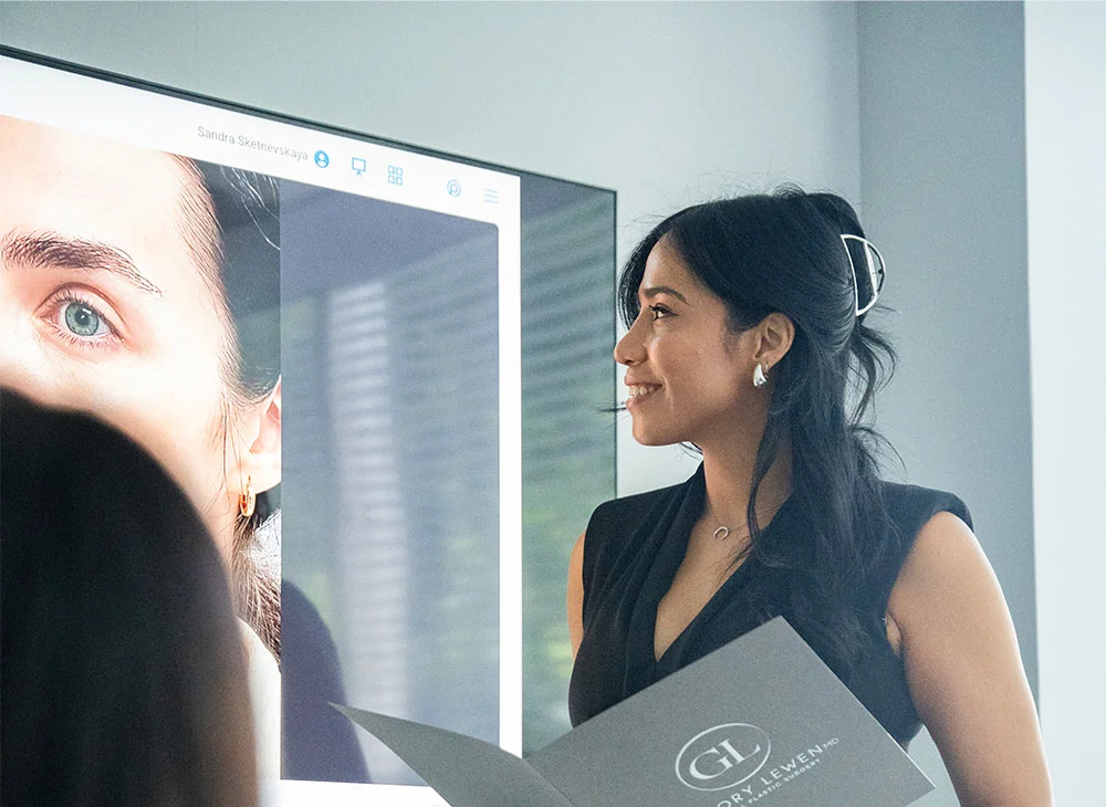 Close up shot of two professionals during a presentation, with one holding a folder and facing a screen. They are conducting a consultation, showing the client's faces on the big screen as part of the discussion. - Tear through Fillers Rejuvenation in Aventura, FL